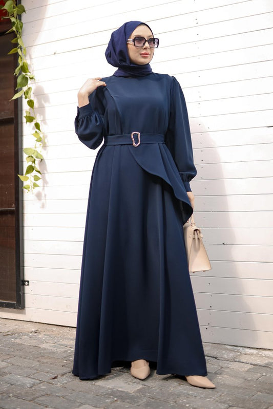 Woman in a navy blue dress with a hijab standing against a white wall.